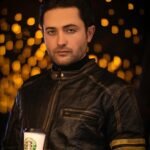A stylish man in a leather jacket holding a coffee cup with warm bokeh lighting in the background.